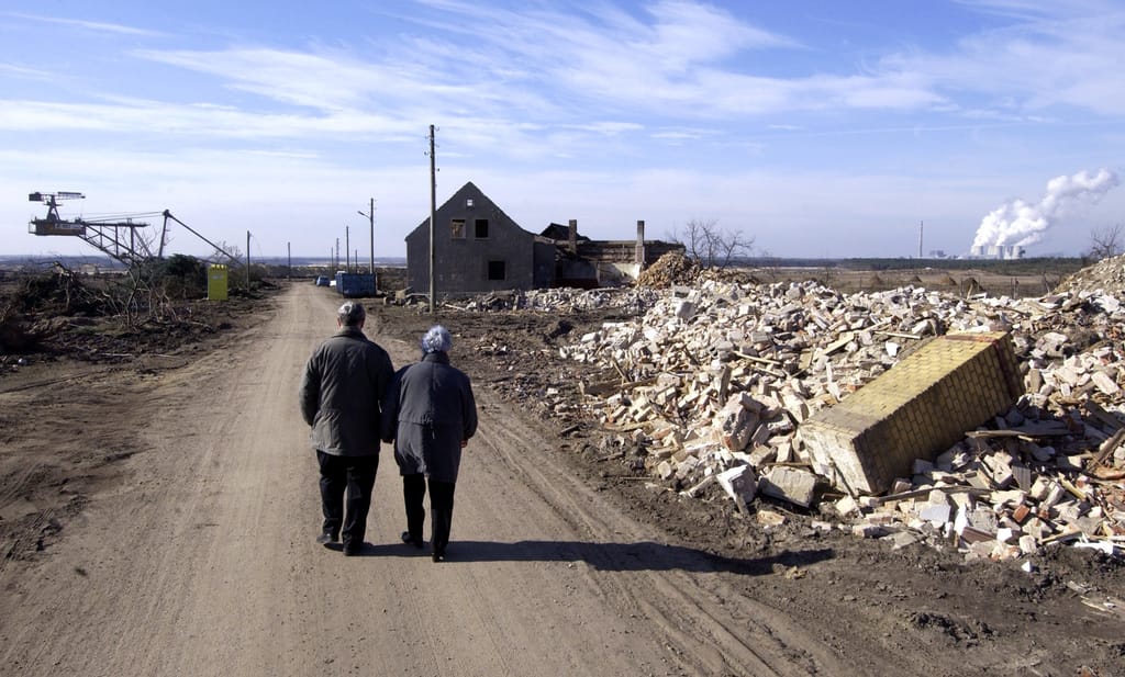 Ein Mann und eine Frau laufen auf an eingerissenen Häusern vorbei und auf ein einzelnes, noch stehendes Haus zu. Noch weiter im Hintergrund sind ein Kohle-Bagger und ein Kraftwerk zu sehen.