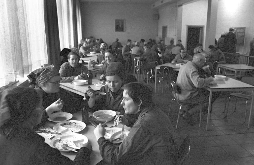 Frauen und Männer sitzen gemeinsam an mehreren einzelnen Tischen beim Mittagessen und Unterhalten sich. Sie tragen alle Arbeitsuniformen.