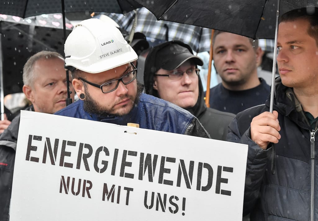 Eine Gruppe Männer steht im Regen. Die meisten halten Regenschirme und im Vordergrund steht ein Mann mit Arbeiterhelm und einem Schild mit der Aufschrift Energiewende nur mit uns.