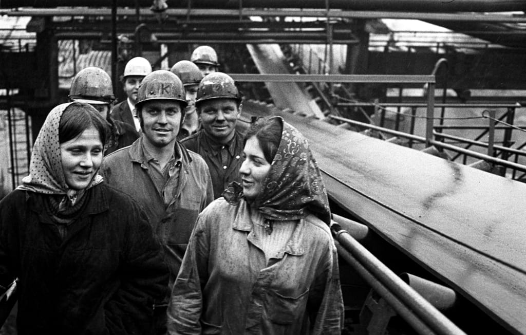 Frauen mit Kopftüchern und Männer mit Helmen stehen in Uniformen neben einem Förderband. Im Hintergrund sind weitere Teile des Kraftwerks zu sehen.