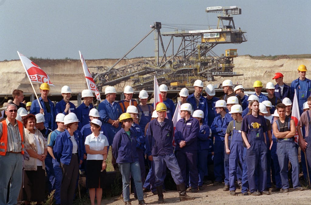Arbeiter und Arbeiterinnen stehen vor einem Tagebau. Sie tragen alle Arbeitsuniformen und halten teilweise Fahnen.